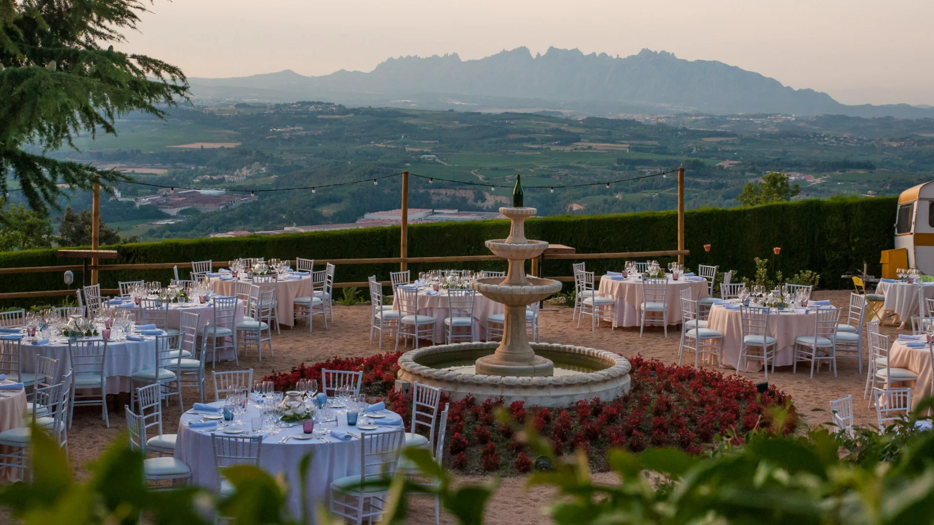 A private outdoor dinner around a stone fountain at a Catalan winery estate, with Montserrat mountain on the horizon at dusk — the kind of setting that earns its place in a multi-venue programme because it has one clear purpose: giving guests an immediate sense of place before the main event begins.
