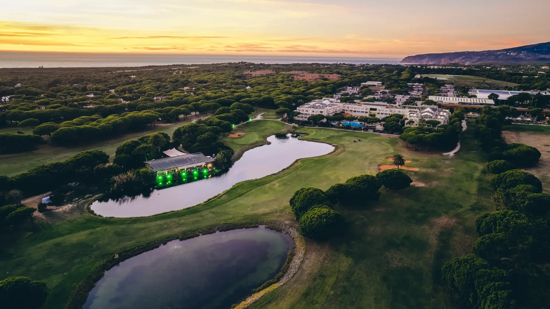 Aerial view at sunset of a golf resort on the Atlantic coast of Portugal, with umbrella pine forests, a lakeside event pavilion and the ocean on the horizon — a destination that earns its place on a retreat shortlist because it fits the brief for space, calm and direct international access, not because it appeared on a wish list first.