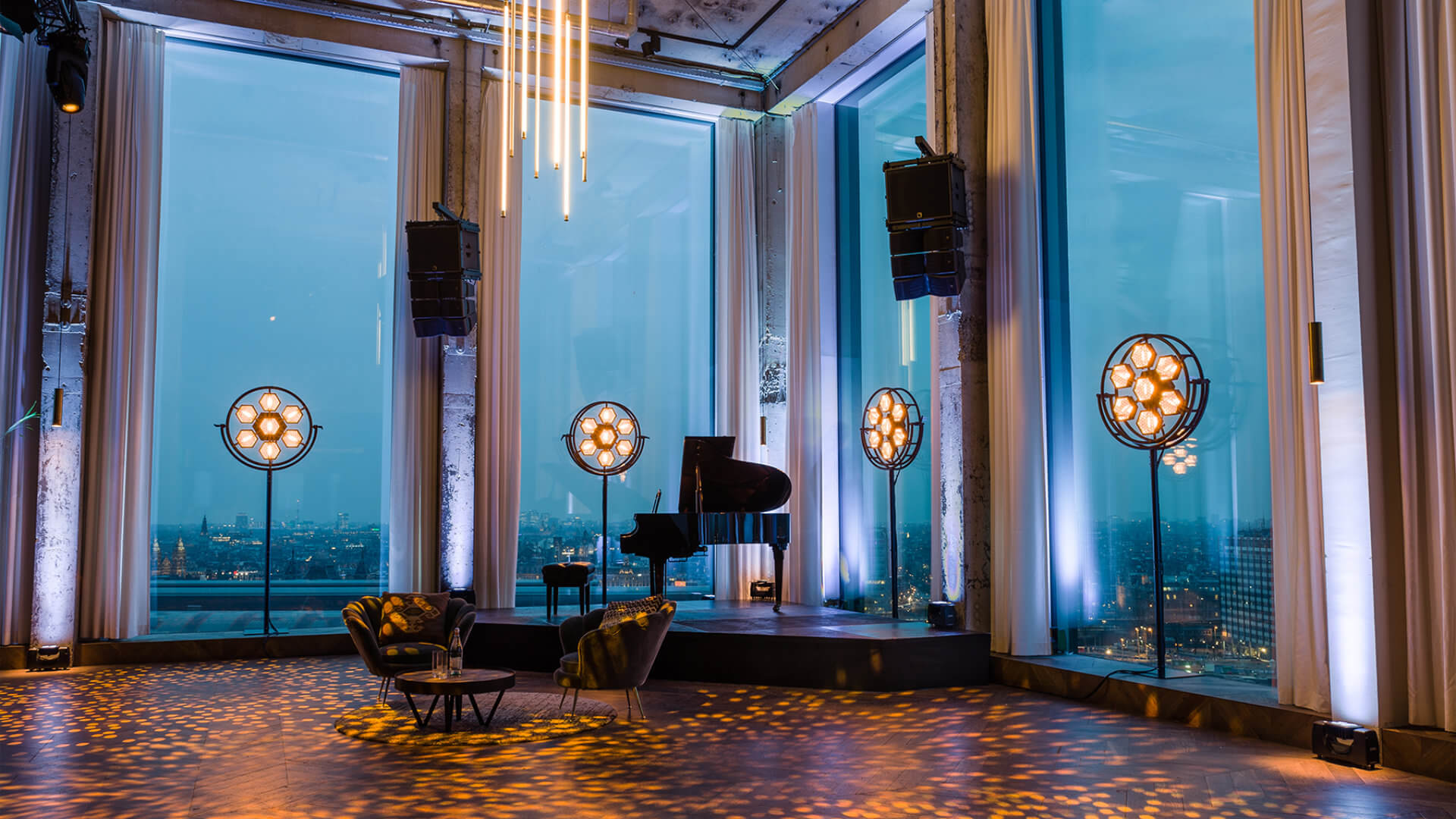 Contemporary Amsterdam event space with floor-to-ceiling windows overlooking the city skyline at dusk, set with a grand piano and decorative globe lights — a contemporary closing venue where an executive dinner can develop into cocktails and music