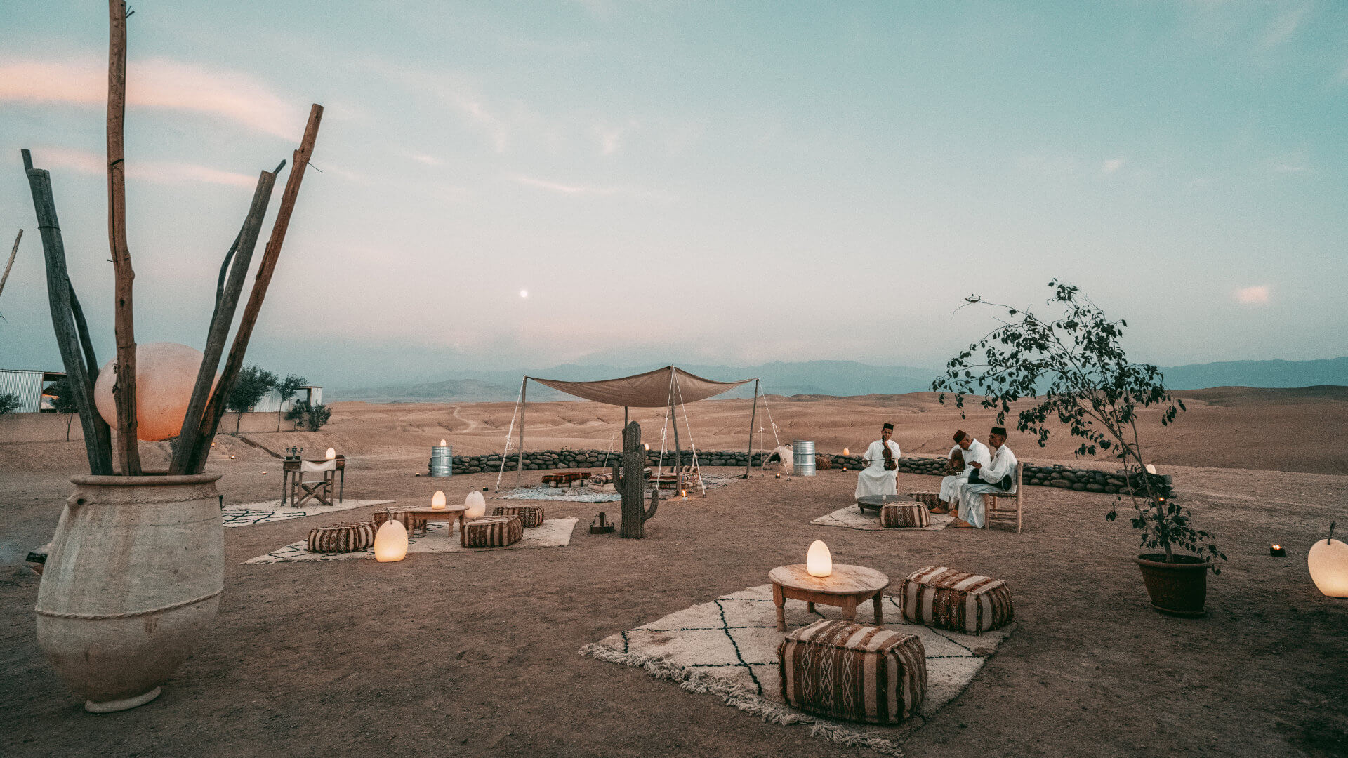 Open-air Agafay desert lounge at dusk with Moroccan ottomans, kilim rugs, glowing lanterns and traditional musicians — an Agafay evening experience that must be planned from the return journey backwards, not the dinner scene forwards