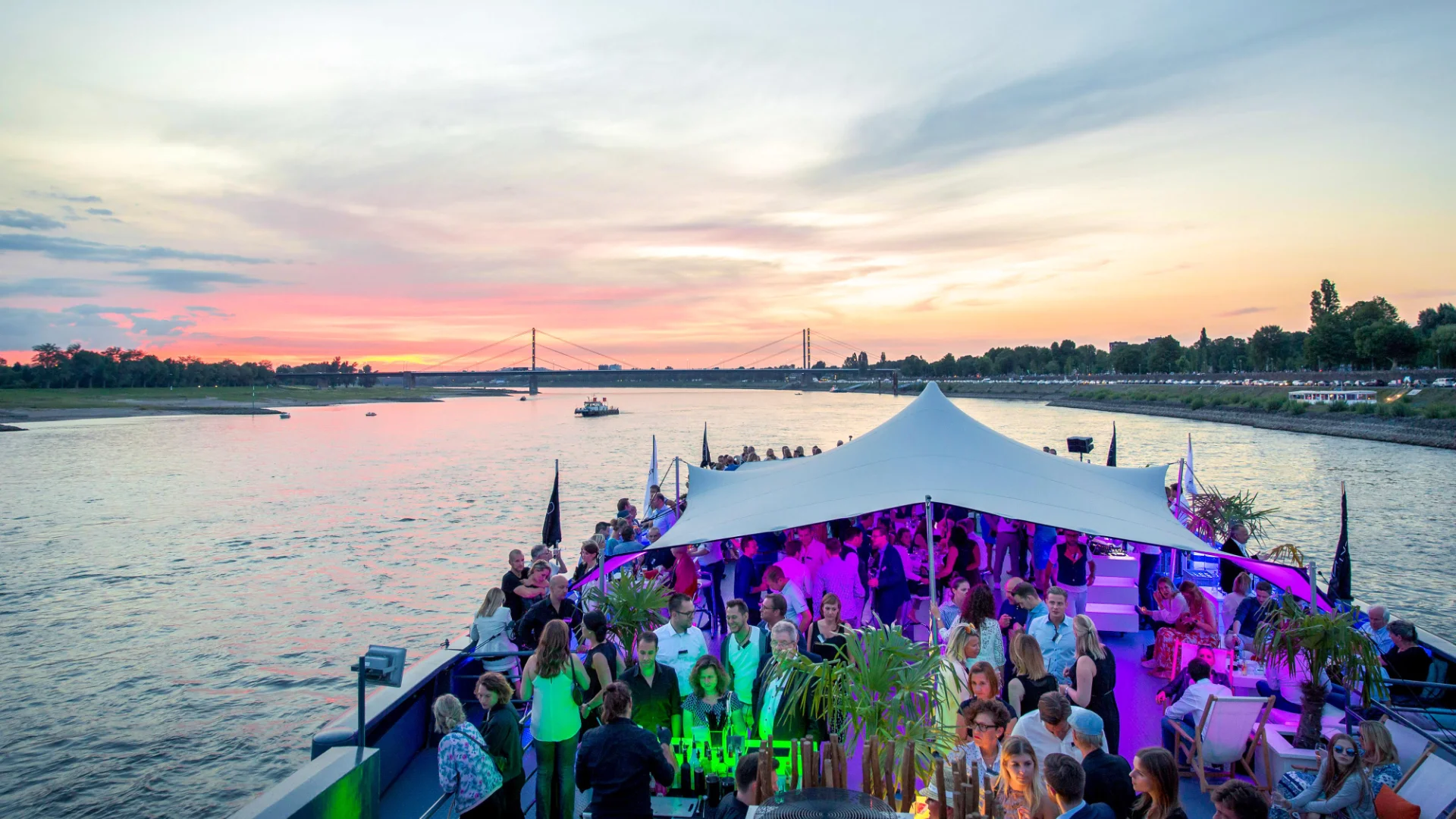 Corporate incentive group celebrating on a river boat at sunset with neon lighting and a crowd under a white canopy tent, the kind of closing event on water that gives a global team space to reconnect before flying home